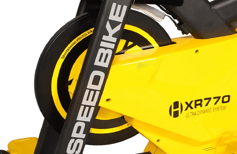 Zbliżenie na żółty rower spinningowy z widocznym napisem \'SPEED BIKE\' na ramie oraz \'XR770 ULTRA DYNAMIC SYSTEM\' na obudowie. Na kole znajduje się tekst \'Spontaneous electric bike\'.