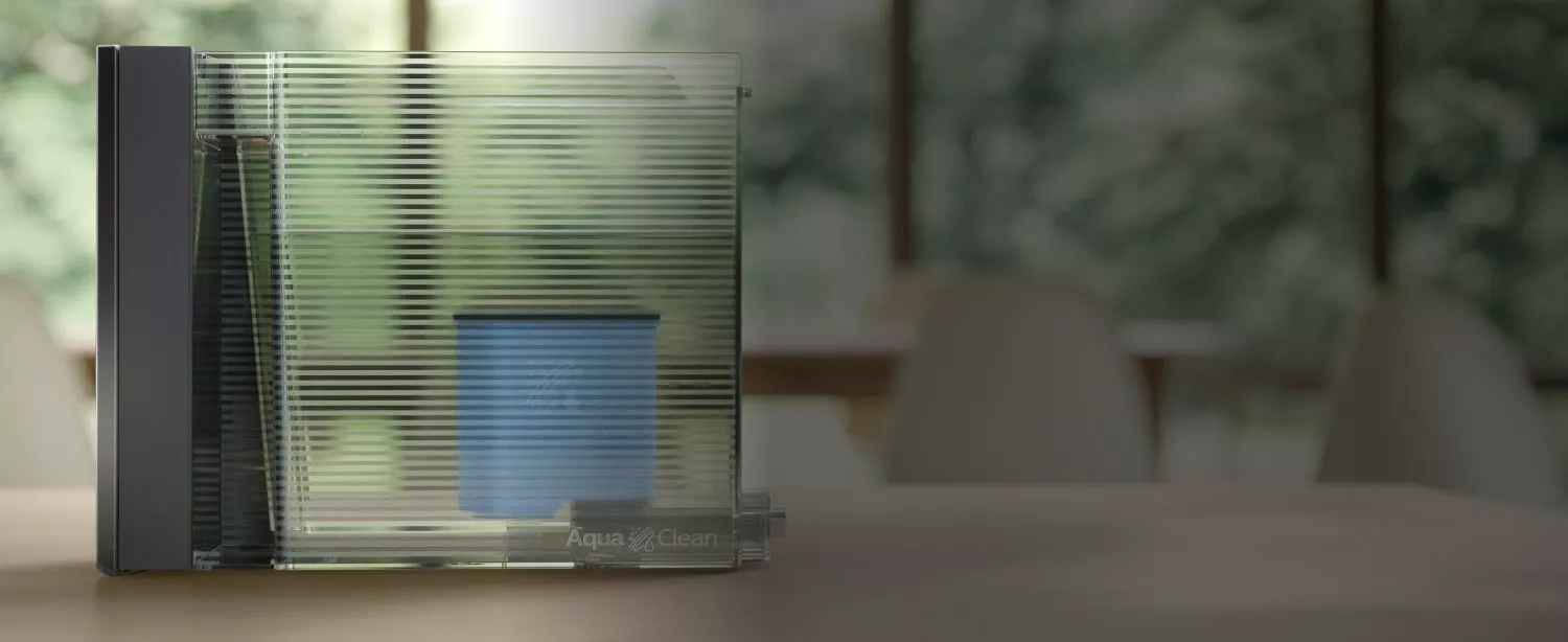Przezroczysty pojemnik z filtrem wody AquaClean, umieszczony na blacie, z widocznymi poziomymi liniami na powierzchni. W tle rozmyty widok wnętrza z krzesłami i oknem.