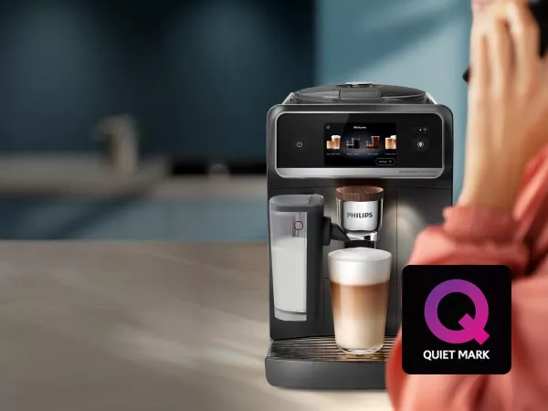 Ekspres do kawy z logo PHILIPS, obok którego znajduje się szklanka z kawą i mlekiem. Na obrazie widoczny jest również symbol \'Q QUIET MARK\'.