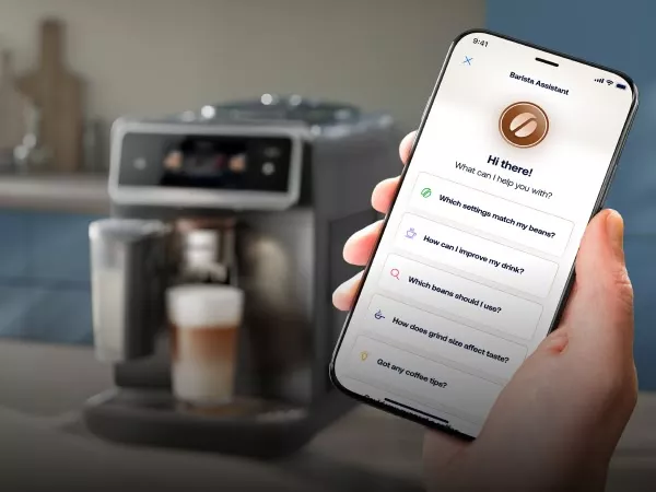 Na pierwszym planie widoczny jest smartfon z aplikacją Barista Assistant, na którym wyświetlane są pytania dotyczące ustawień kawy. W tle znajduje się ekspres do kawy z przygotowanym napojem w szklance.