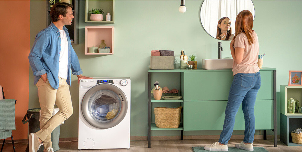W jasnym pomieszczeniu znajduje się pralka z widocznym logo \'Candy\', obok której stoi mężczyzna. Kobieta stoi przy umywalce, patrząc w lustro, a na półkach znajdują się różne dekoracje i rośliny.
