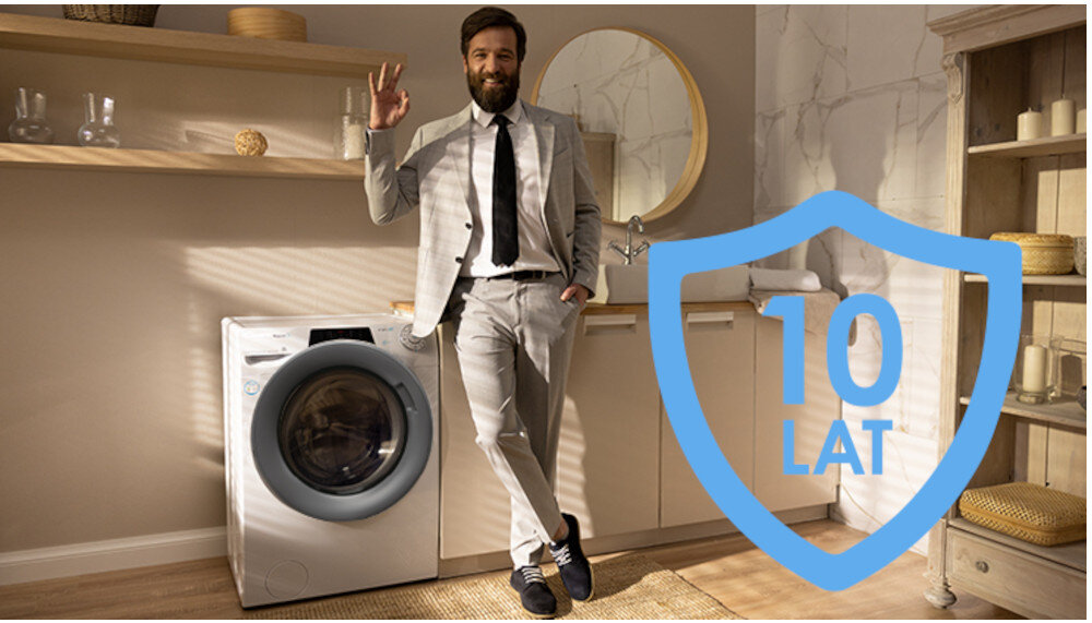 Mężczyzna w garniturze stoi obok pralki w jasnym pomieszczeniu. Na obrazie widoczny jest niebieski symbol z napisem \'10 LAT\'.