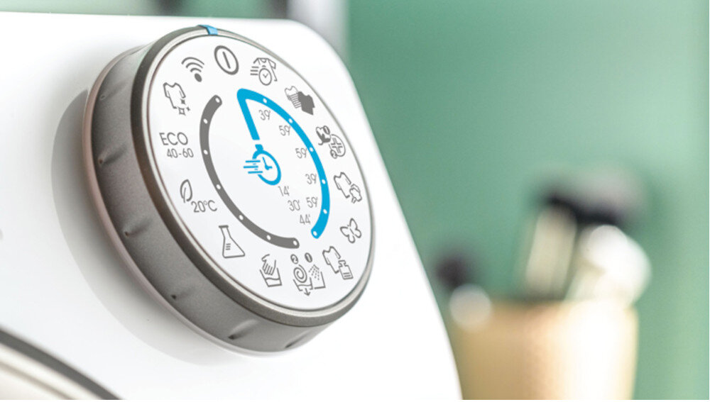 Zbliżenie na pokrętło pralki z różnymi ikonami programów prania, w tym ECO 40-60 i 20°C. Pokrętło jest umieszczone na białej powierzchni, a w tle widać rozmyte elementy wnętrza.