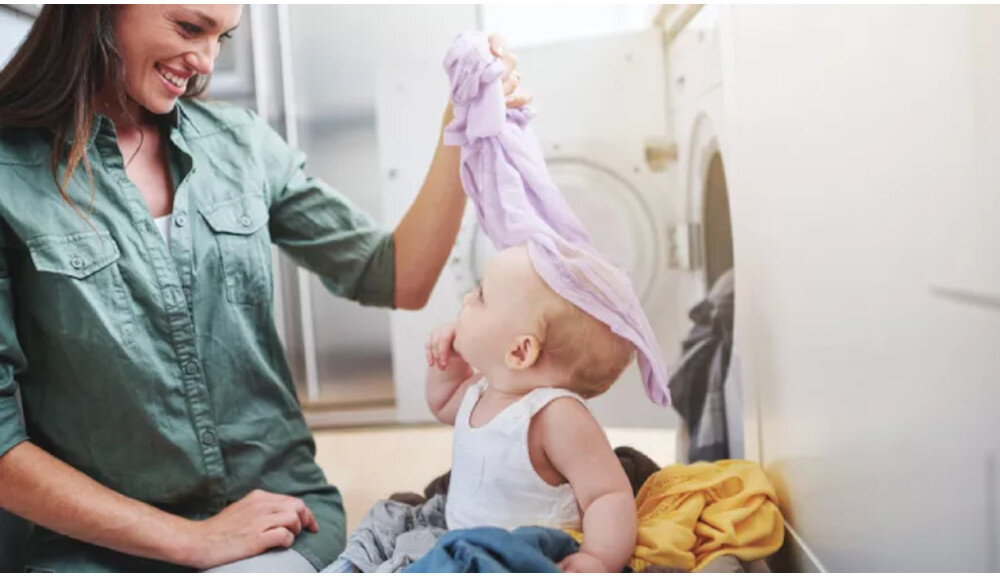 Kobieta i dziecko siedzą obok pralki, kobieta trzyma w ręku kawałek tkaniny. W tle widoczna jest otwarta pralka z bębnem.