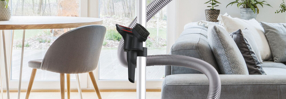 Wnętrze salonu z widocznym stołem, krzesłem i sofą. Na pierwszym planie znajduje się rura odkurzacza z przymocowaną szczotką.