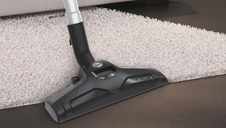 Na obrazie widoczna jest końcówka odkurzacza na granicy dywanu i podłogi. Na końcówce znajduje się logo \'Hoover\'.