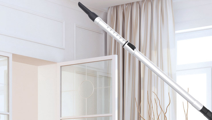 Na obrazie widoczna jest metalowa rura odkurzacza z czarną końcówką, umieszczona na tle jasnego wnętrza z zasłonami i otwartym oknem.