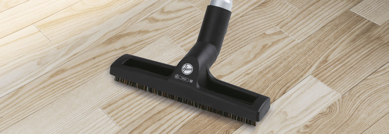 Na obrazie widoczna jest czarna szczotka odkurzacza na drewnianej podłodze. Na szczotce znajduje się logo z napisem \'Hoover\'.