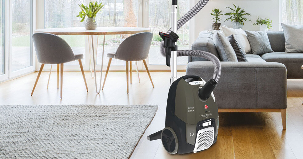 Odkurzacz w kolorze ciemnym z widocznym logo i napisem \'HOOVER\' oraz \'TELIOS PLUS\' stoi na drewnianej podłodze w salonie. W tle znajduje się szara sofa, stolik z krzesłami i duże okna z widokiem na ogród.