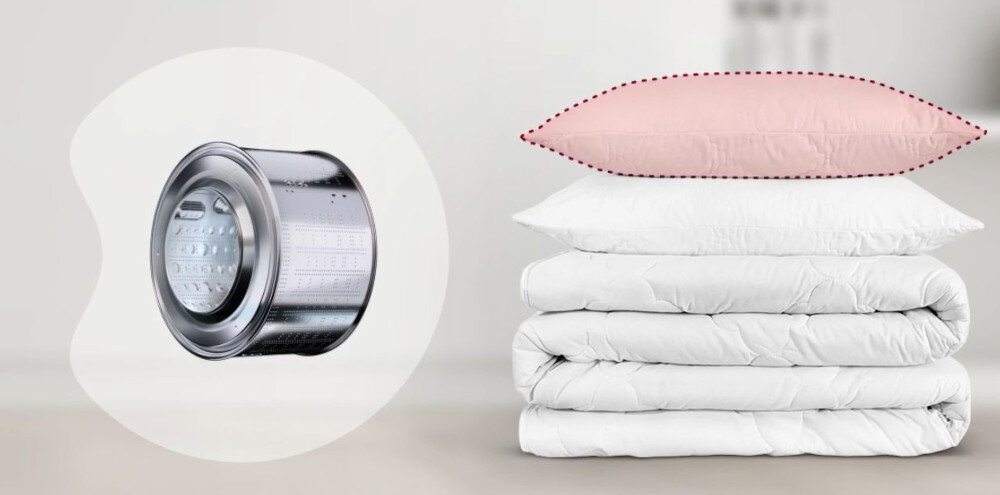 Obraz przedstawia bęben pralki oraz stos białych poduszek z różową poduszką na górze. Bęben pralki jest metaliczny i błyszczący, a poduszki są ułożone na sobie na jasnym tle.