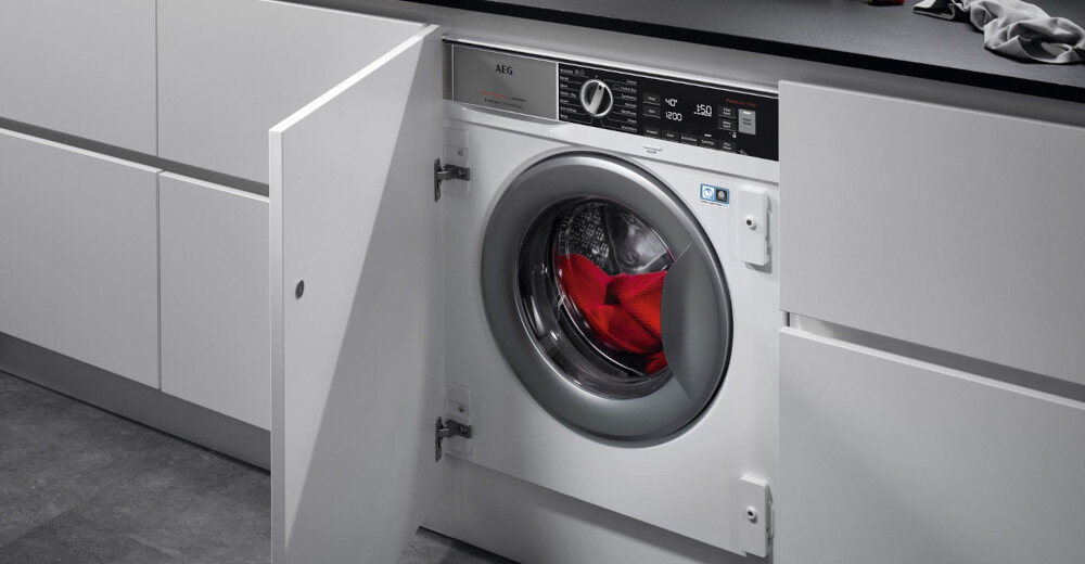 Pralka w zabudowie z otwartymi drzwiami, w bębnie widoczne czerwone ubranie. Na panelu sterowania widoczny jest napis \'AEG\' oraz pokrętło i przyciski.