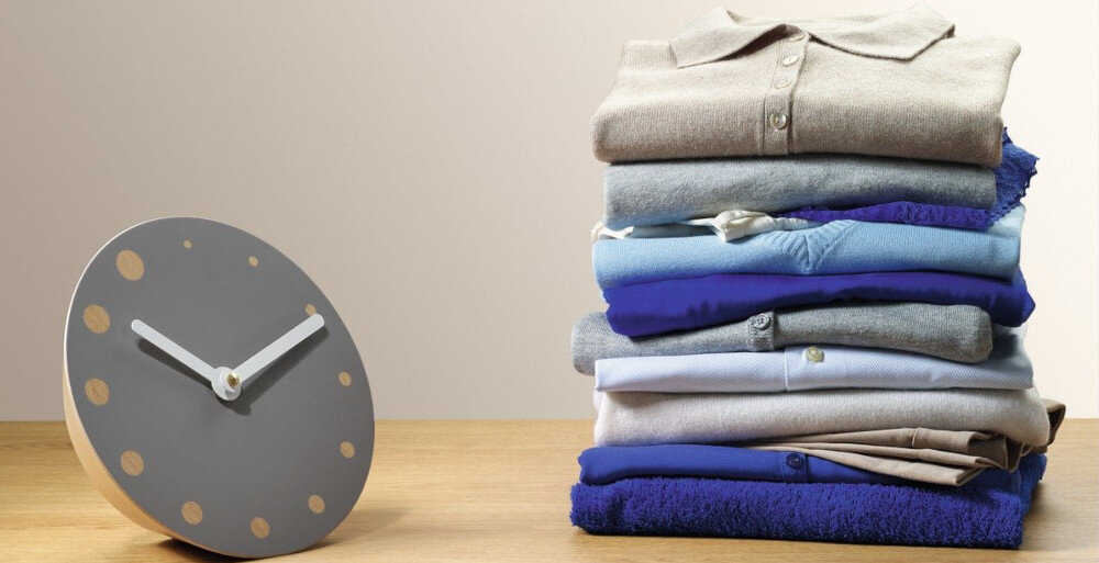 Na drewnianym blacie stoi szary zegar z białymi wskazówkami i złotymi kropkami zamiast cyfr. Obok znajduje się stos złożonych ubrań w różnych odcieniach niebieskiego i beżu.