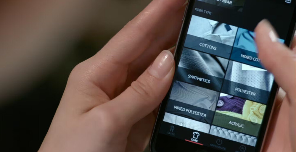Osoba trzyma smartfon z aplikacją pokazującą różne typy tkanin, takie jak \'COTTONS\', \'MIXED COTTON\', \'SYNTHETICS\', \'POLYESTER\', \'MIXED POLYESTER\', \'ACRYLIC\'.