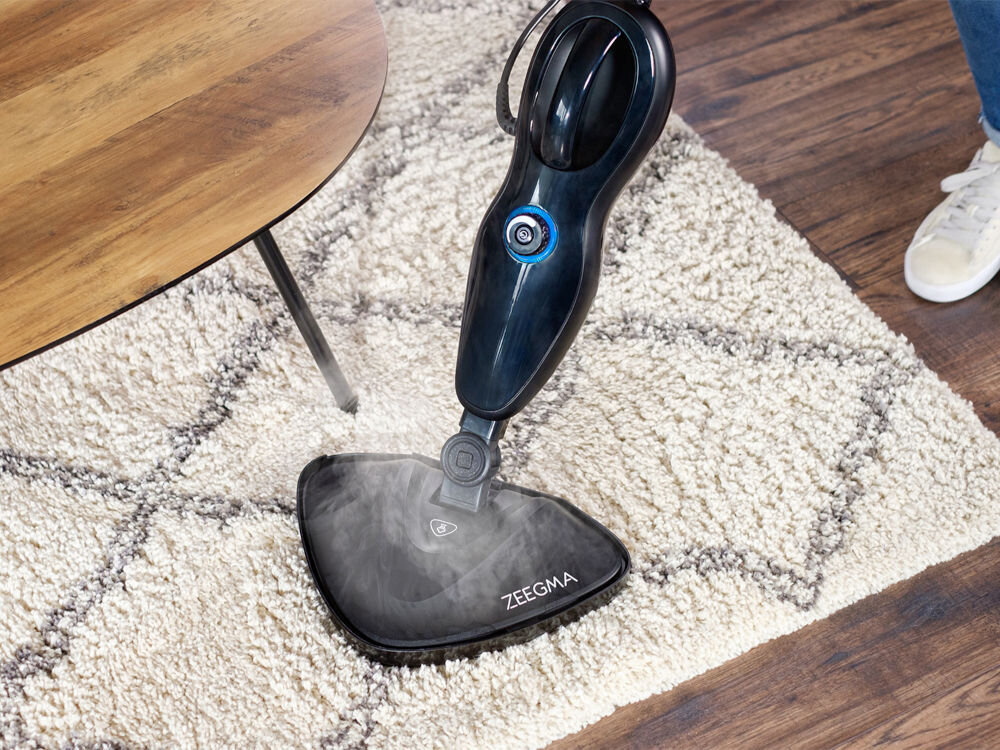 Mop parowy marki ZEEGMA jest używany na dywanie, wydzielając parę. Obok znajduje się drewniany stolik i fragment buta osoby obsługującej urządzenie.