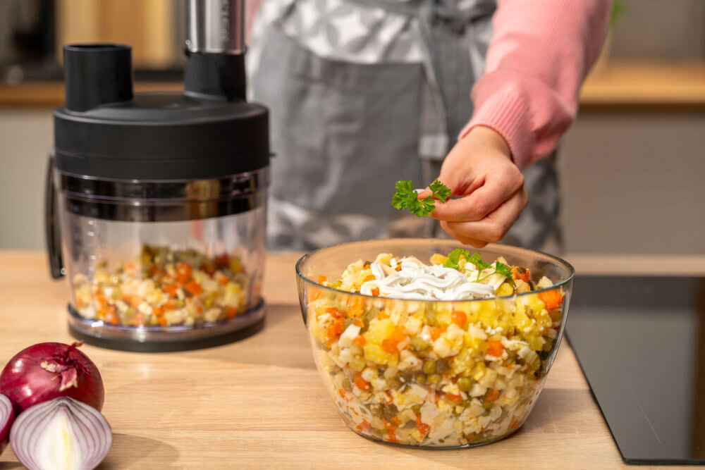 Na blacie kuchennym stoi blender z pojemnikiem wypełnionym pokrojonymi warzywami. Obok znajduje się miska z sałatką, a osoba dodaje do niej gałązkę natki pietruszki.