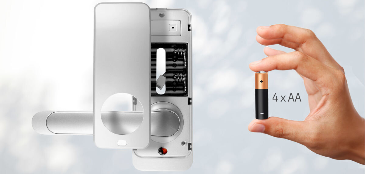 Obraz przedstawia srebrną klamkę z otwartą pokrywą, ukazującą miejsce na baterie. Obok widoczna jest ręka trzymająca baterię AA z napisem \'4 x AA\'.