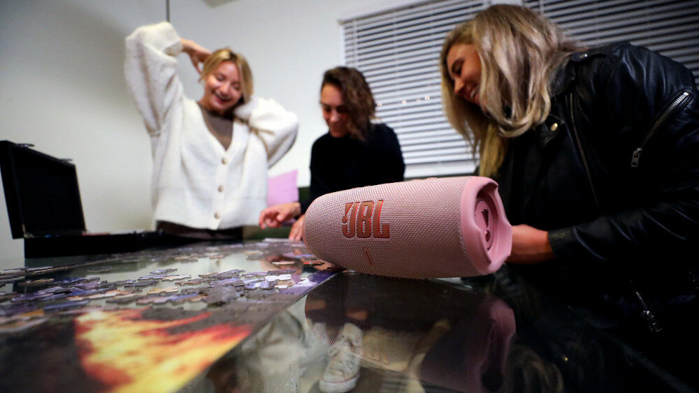 Na stole leży różowy głośnik z widocznym logo \'JBL\'. W tle znajdują się trzy osoby, które układają puzzle.