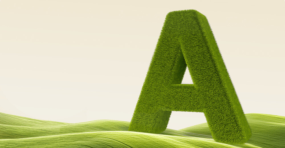 Duża litera \'A\' pokryta zieloną trawą stoi na tle zielonych, falujących wzgórz. Tło jest jasne, co podkreśla kontrast z zielenią litery i wzgórz.