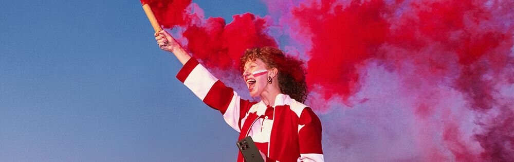 Osoba ubrana w biało-czerwony strój trzyma w ręku czerwoną racę dymną, a w tle unosi się czerwony dym na tle nieba.