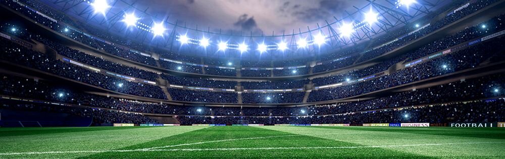 Oświetlony stadion piłkarski z zieloną murawą, otoczony trybunami pełnymi widzów. Na bandach reklamowych widoczne są różne logotypy i napisy, w tym \'FOOTBALL\'.