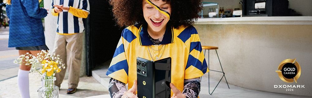 Osoba siedzi przy stoliku na zewnątrz, trzymając w rękach składany smartfon. W tle widoczne są inne osoby oraz logo \'DXOMARK 2026\' z napisem \'GOLD CAMERA\'.