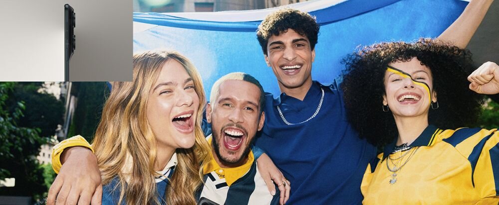 Grupa czterech osób uśmiecha się i trzyma flagę w tle. W lewym górnym rogu widoczny jest bok smartfona.