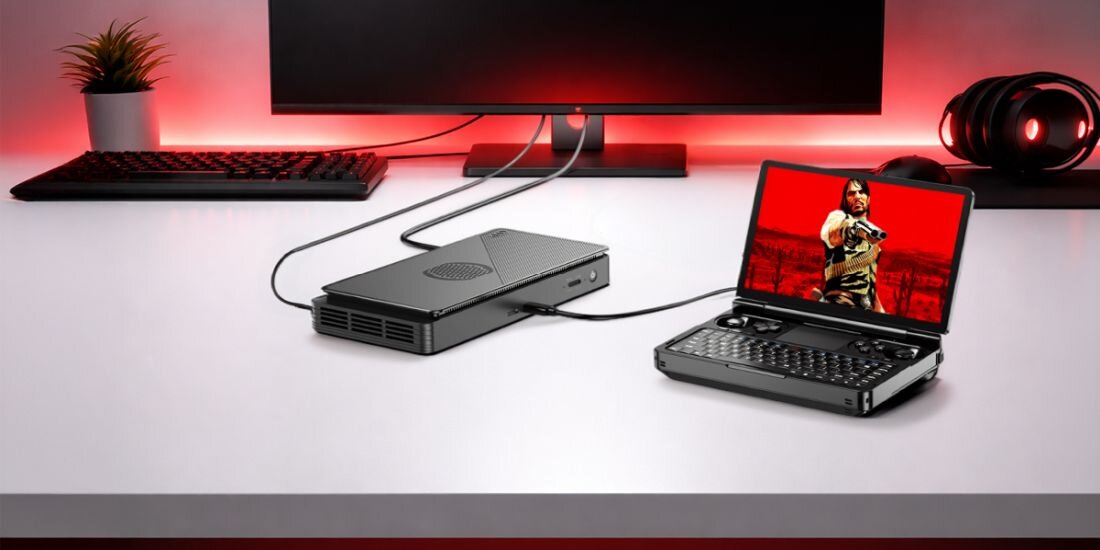 Na białym biurku znajduje się otwarty laptop z wyświetlonym obrazem postaci oraz podłączona do niego czarna konsola. W tle widoczny jest monitor, słuchawki i czerwone podświetlenie.