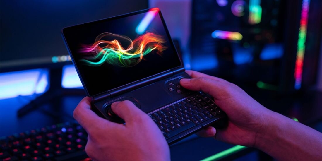 Osoba trzyma mały laptop z otwartym ekranem, na którym widoczna jest kolorowa grafika z falami. W tle znajduje się klawiatura i komputer z podświetleniem RGB.
