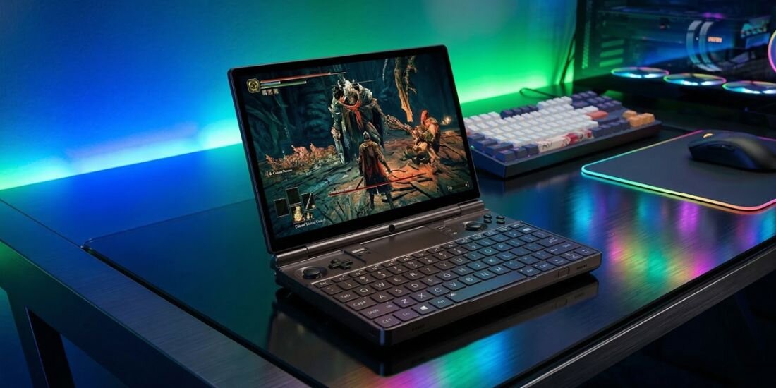 Na biurku znajduje się otwarty laptop z wyświetlonym na ekranie obrazem z gry. Obok laptopa leży klawiatura i myszka na podświetlanej podkładce.