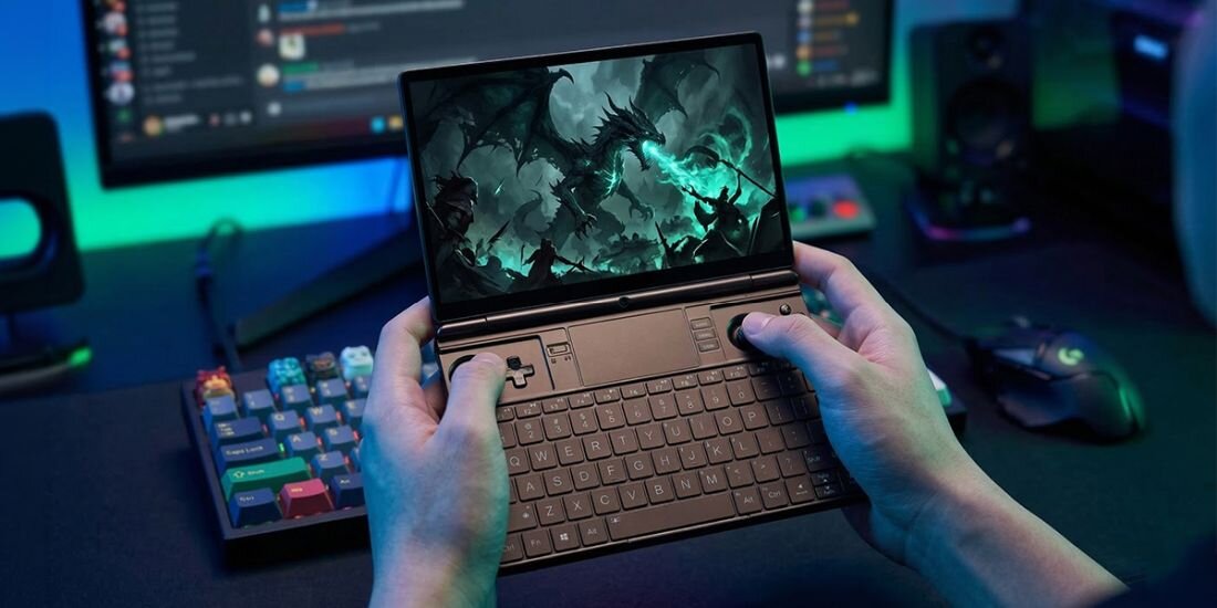 Osoba trzyma mały laptop z otwartym ekranem, na którym wyświetlana jest gra z motywem smoków. W tle widać klawiaturę, myszkę i monitor na biurku.