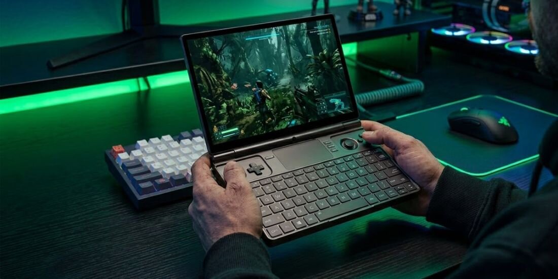 Osoba trzyma laptopa z otwartym ekranem, na którym wyświetlana jest gra w scenerii dżungli. Na biurku obok znajduje się klawiatura i myszka, a w tle widać podświetlenie LED.
