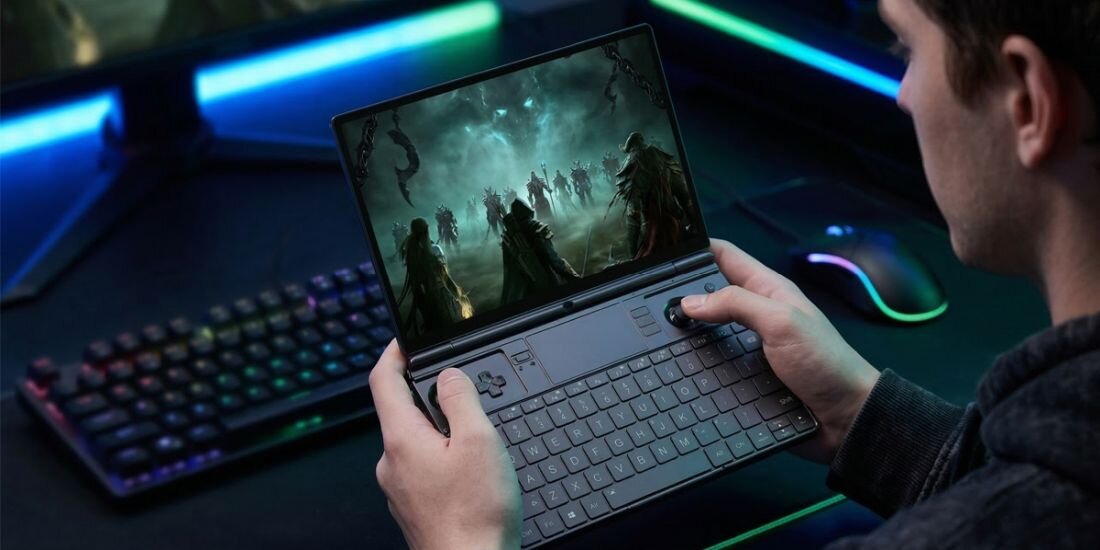 Osoba trzyma laptop z otwartym ekranem, na którym wyświetlana jest gra z postaciami w mrocznej scenerii. Na klawiaturze laptopa widoczne są klawisze i touchpad.