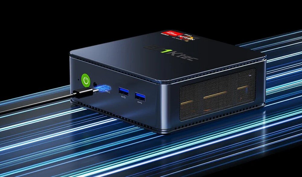 Na obrazie widoczny jest kompaktowy komputer z logo \'GMKTEC\' na górnej powierzchni. Z przodu znajdują się dwa porty USB, przycisk zasilania oraz podłączony kabel.