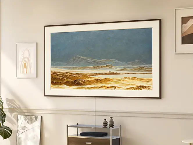 2026-ls03ha-top-4-modern-frame-design-with-one-connect 664x498 Na ścianie wisi telewizor z wyświetlonym obrazem przedstawiającym krajobraz górski z niebieskim niebem i złotymi wzgórzami. Pod telewizorem znajduje się półka z dekoracjami.