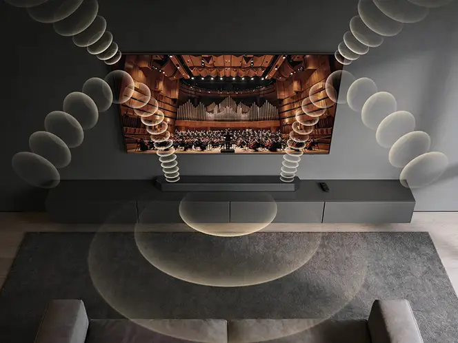 2026-dynamic-sound-pack-2-q-symphony 664x498 Na ścianie wisi duży telewizor wyświetlający obraz sali koncertowej z orkiestrą. Wokół telewizora widoczne są graficzne fale dźwiękowe, a poniżej znajduje się szafka z pilotem.