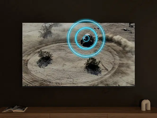Telewizor wyświetla obraz pustynnego krajobrazu z pojazdem terenowym, wokół którego widoczne są niebieskie okręgi. Na półce pod telewizorem znajduje się mała dekoracja.