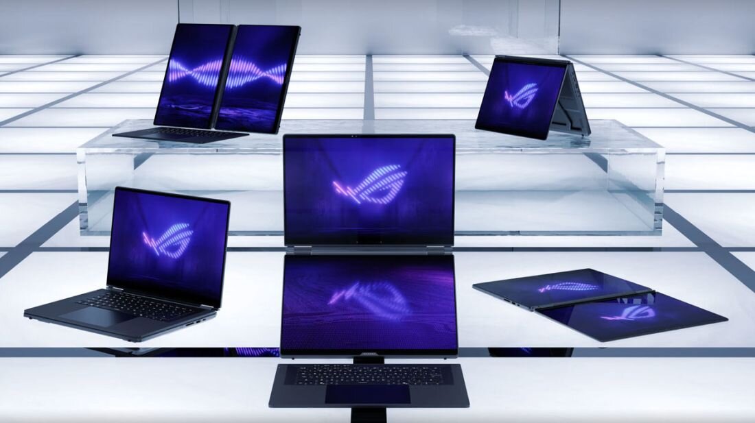 Na obrazie widoczne są laptopy z logo ROG na ekranach, ustawione na szklanej powierzchni w jasnym, nowoczesnym wnętrzu. Ekrany laptopów wyświetlają fioletowy wzór przypominający falę.