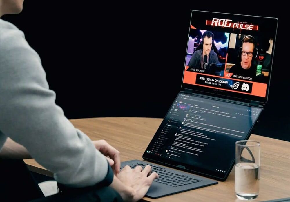 Osoba korzysta z laptopa z dwoma ekranami, na górnym ekranie widoczny jest program \'ROG Pulse\' z dwoma mężczyznami i tekstem \'JOIN US ON DISCORD\'. Obok laptopa stoi szklanka z wodą na drewnianym stole.