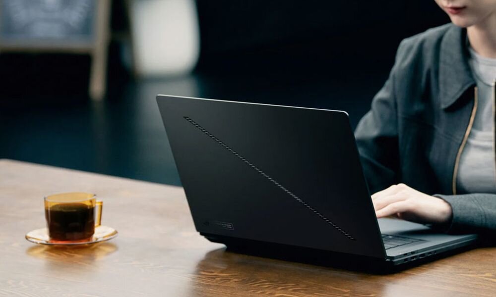 Osoba korzysta z czarnego laptopa na drewnianym stole, obok stoi szklanka z napojem. Laptop ma minimalistyczny design z subtelnym wzorem na obudowie.