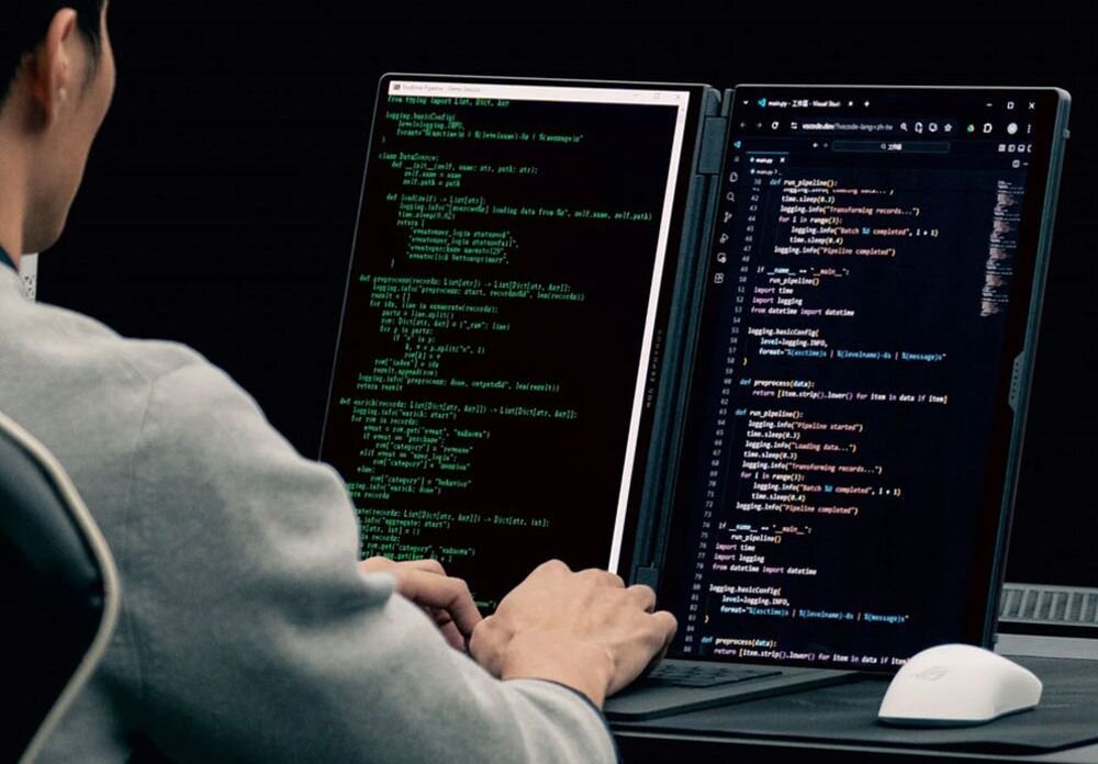 Osoba pracuje na laptopie z dwoma ekranami, na których widoczny jest kod programistyczny. Obok laptopa znajduje się mysz komputerowa.