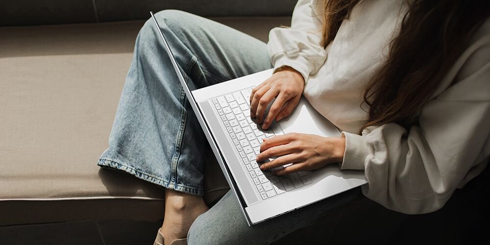 Osoba siedzi na kanapie i używa laptopa z białą klawiaturą. Laptop jest otwarty, a dłonie osoby spoczywają na klawiaturze.