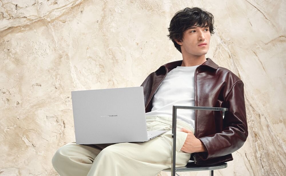 Osoba siedzi na krześle, trzymając na kolanach otwarty laptop z widocznym logo \'ASUS Zenbook\'. W tle znajduje się jasna, marmurowa ściana.