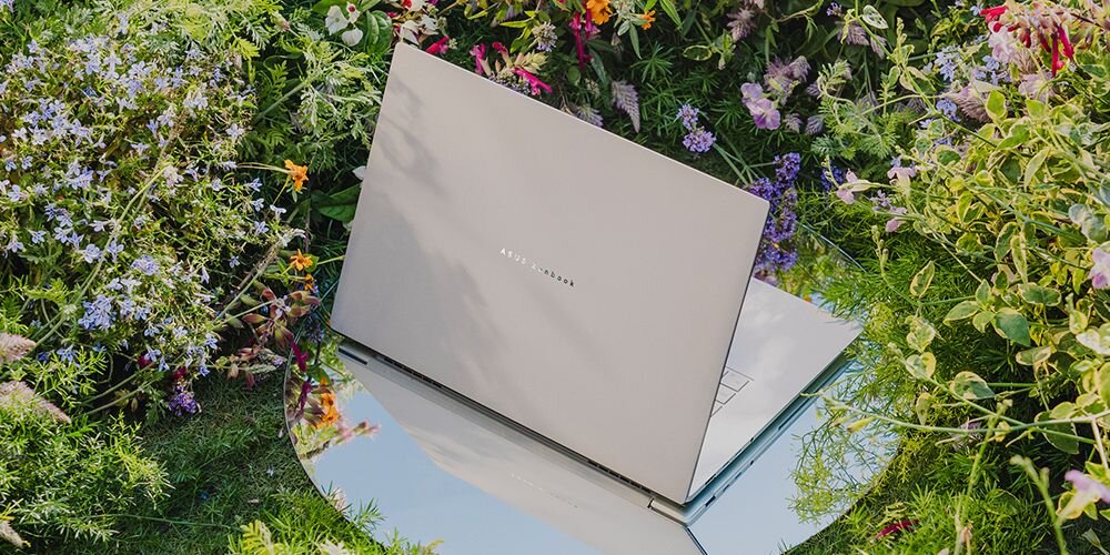 Laptop z widocznym logo \'ASUS Zenbook\' stoi na lustrzanej powierzchni otoczonej roślinnością i kwiatami.