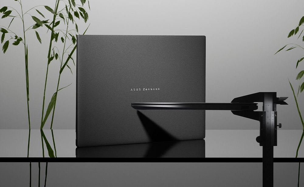 Na obrazie widać zamknięty laptop z napisem \'ASUS Zenbook\' na pokrywie. W tle znajdują się zielone rośliny, a obok laptopa widoczny jest czarny element konstrukcyjny.