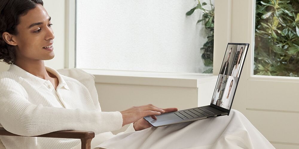 Osoba siedzi na kanapie, trzymając laptopa na kolanach. Na ekranie laptopa widoczny jest podział na cztery okna z rozmową wideo.