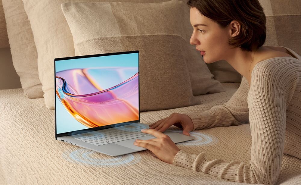 Osoba leży na łóżku i korzysta z laptopa, na ekranie którego widoczna jest abstrakcyjna grafika w jasnych kolorach. Laptop znajduje się na beżowej narzucie, a wokół niego widoczne są efekty świetlne sugerujące dźwięk.