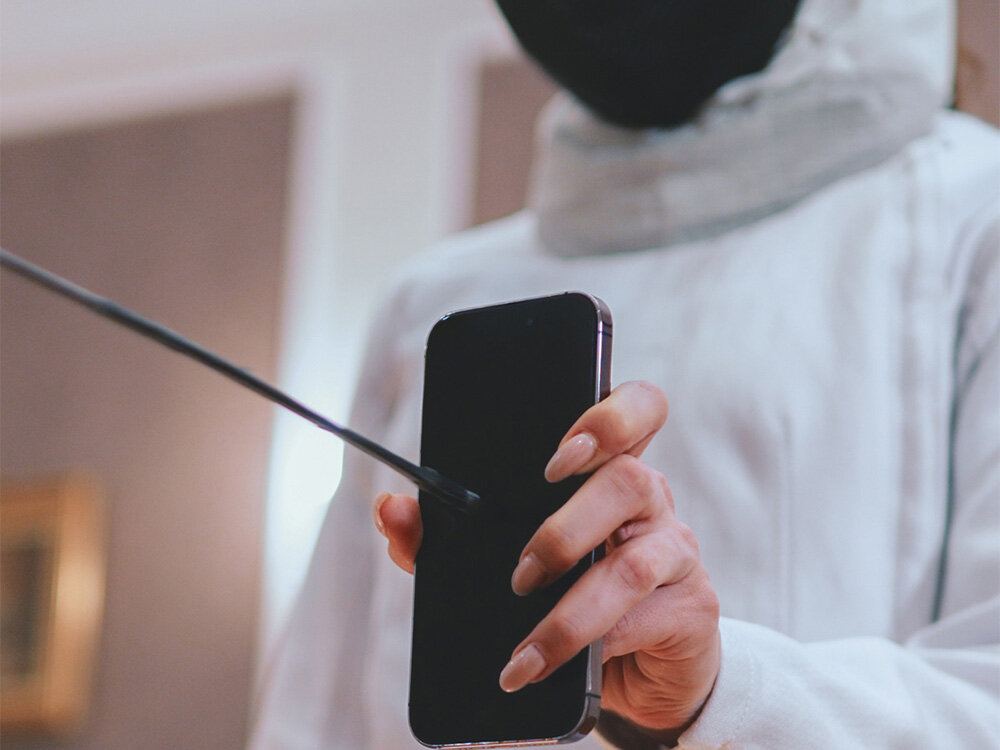 Osoba w białym ubraniu trzyma smartfon, na który wskazuje cienkim przedmiotem. Tło jest rozmyte, a osoba ma na sobie czarną maskę.