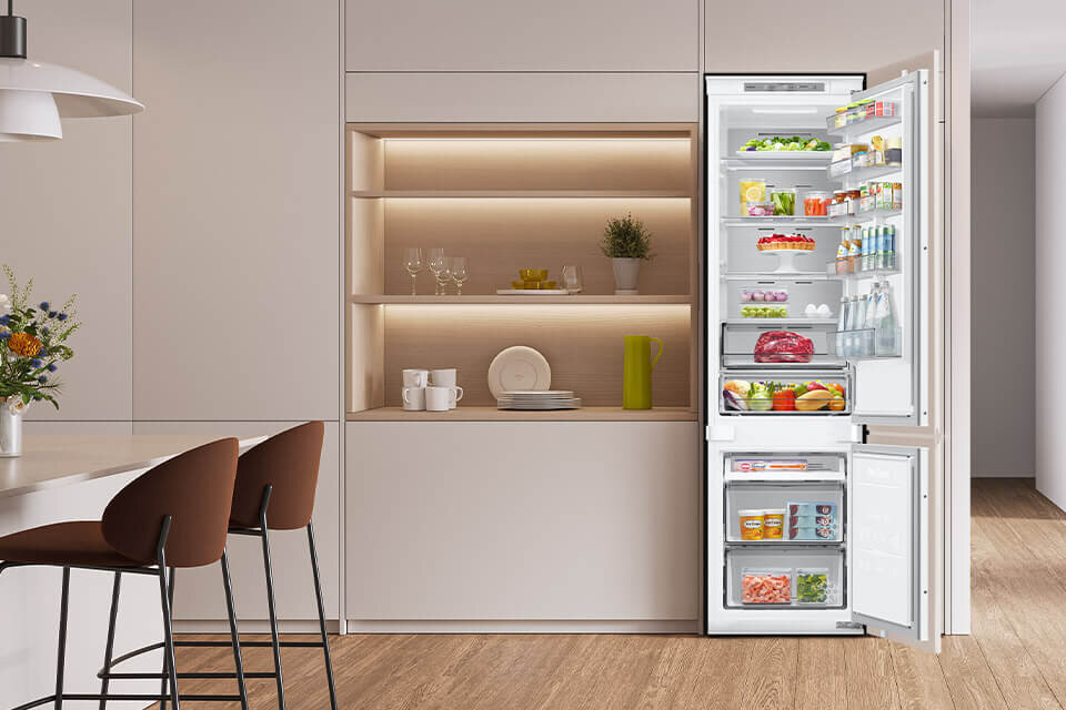 Wnętrze nowoczesnej kuchni z otwartą lodówką w zabudowie, wypełnioną różnorodnymi produktami spożywczymi. Obok lodówki znajduje się półka z naczyniami i dekoracjami, a na blacie stoi wazon z kwiatami.