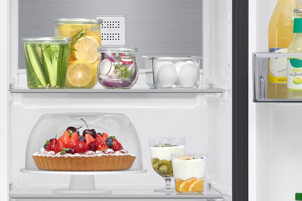 Wnętrze lodówki z półkami, na których znajdują się różne produkty spożywcze, w tym słoiki z warzywami i cytrynami, jajka, ciasto z owocami oraz napoje w butelkach. Na jednej z półek widoczne są również szklanki z deserami z kiwi i pomarańczami.