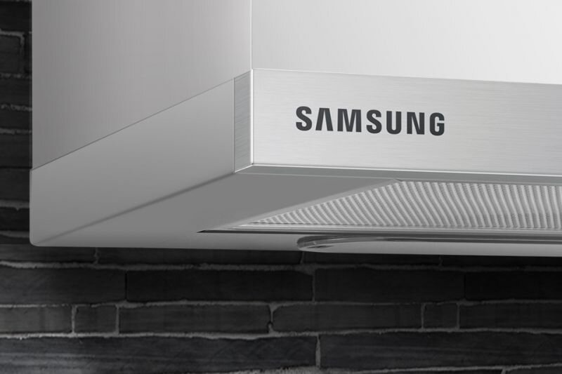 Fragment okapu kuchennego z widocznym logo \'SAMSUNG\' na stalowej powierzchni. W tle znajduje się ściana z ciemnych cegieł.